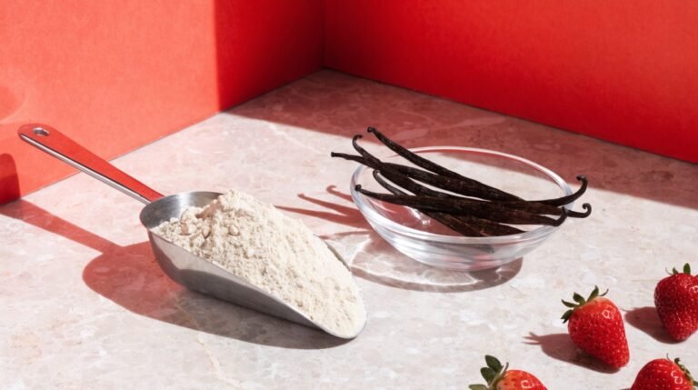 A scoop of unflavored protein powder and a small glass bowl of vanilla pods on a pale stone counter against a bold coral background with fresh strawberries