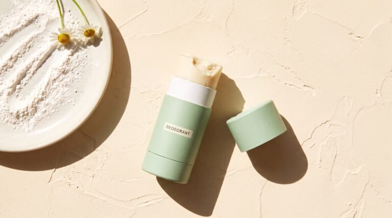 A baking-soda-free natural deodorant stick beside a small dish of white powder and a sprig of chamomile on a pale surface in bright daylight