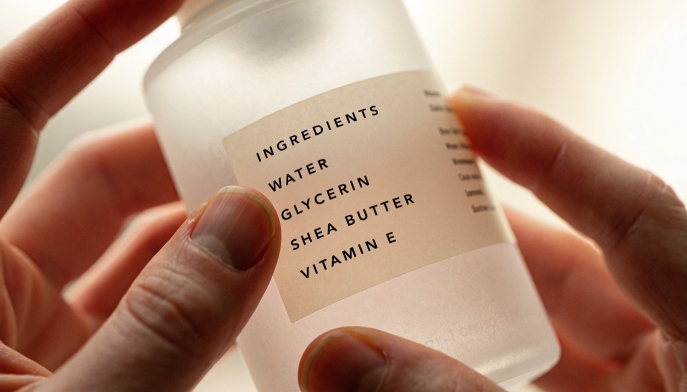 A hand checking the ingredient label on the back of a frosted skincare bottle