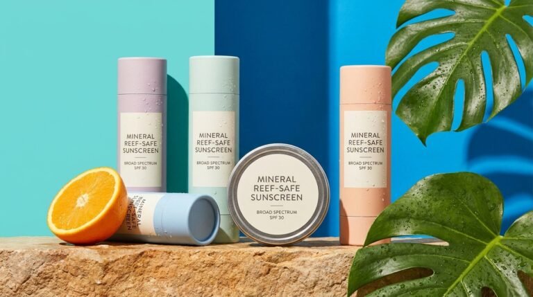 Mineral reef-safe sunscreen tubes and tins on a stone ledge against a bright blue background with tropical leaves and citrus