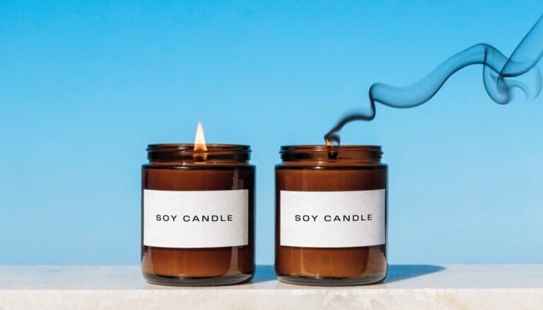 Two amber jar candles labelled SOY CANDLE on a pale stone ledge against a deep sky-blue background, one burning cleanly on a trimmed wick and one smoking from an overlong wick