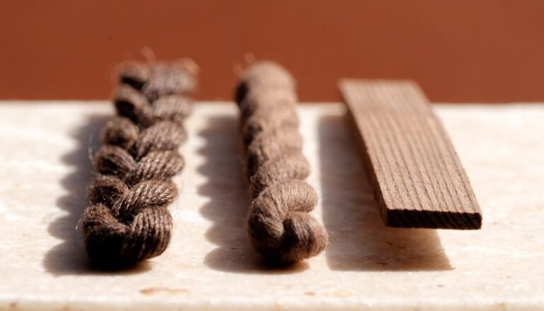 Three unlit candle wicks side by side, flat cotton, braided cotton and a flat wooden wick, on a pale stone ledge against a warm terracotta background