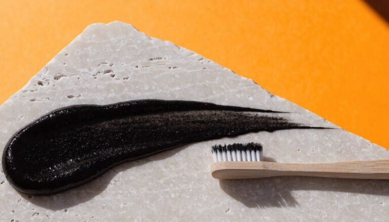 A smear of black charcoal toothpaste pulled across a pale stone slab beside a bamboo toothbrush against a bright citrus-orange background