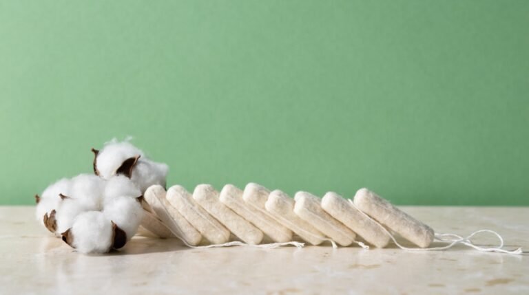A neat row of unbleached cotton tampons beside a small pile of raw cotton on a pale stone surface against a bold sage background in soft daylight