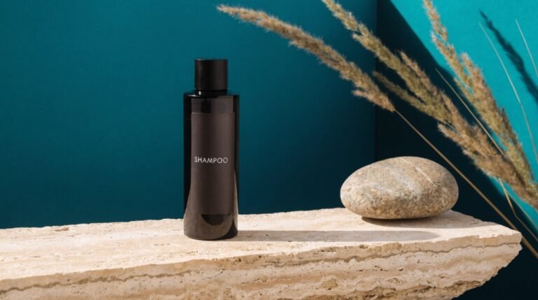 A dark matte bottle labeled Shampoo standing on a travertine ledge against a bold deep-teal background with dried grasses and a smooth river stone
