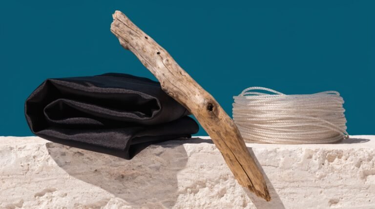 A folded black swimsuit and a coil of fine nylon rope on a pale stone ledge against a bold deep-teal background with a piece of weathered driftwood