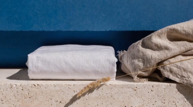A crisply folded white sheet and a linen pillowcase on a pale stone ledge against a bold dusty-blue background with a dried grass stem in soft light