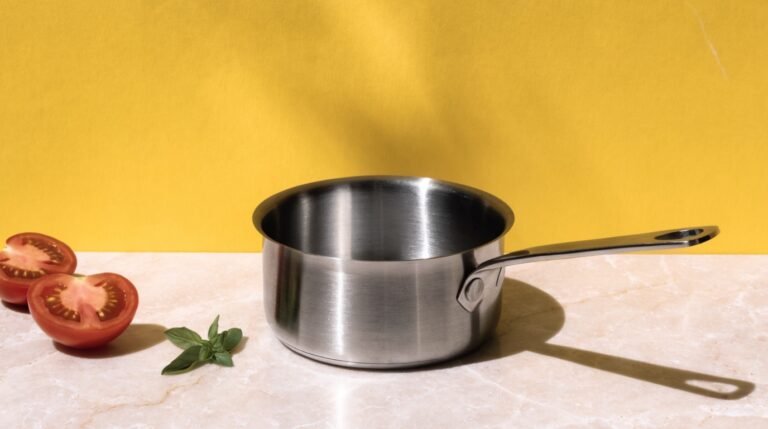 A brushed stainless saucepan on a pale stone counter against a bold mustard-yellow background with two halved tomatoes and a sprig of basil in hard sunlight