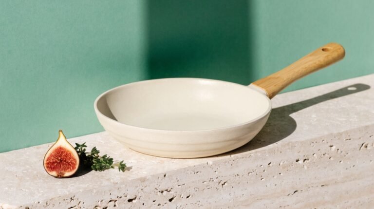 A cream ceramic-coated frying pan with a wooden handle on a travertine ledge against a bold sage-green background with a halved fig and fresh thyme