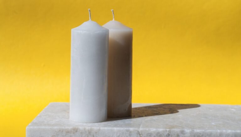 A plain white paraffin pillar candle and a cream soy candle side by side on a pale stone ledge against a bright sunny-yellow background