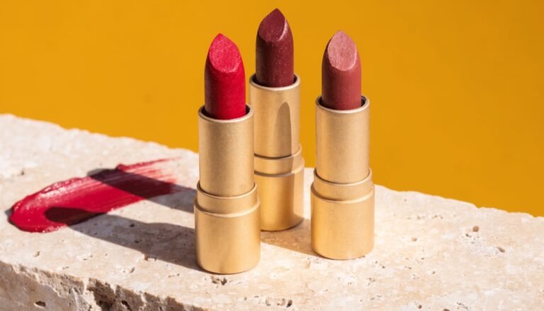 Three open lipstick bullets in deep red and berry shades arranged on a pale stone ledge against a bold mustard-yellow background in bright directional sunlight