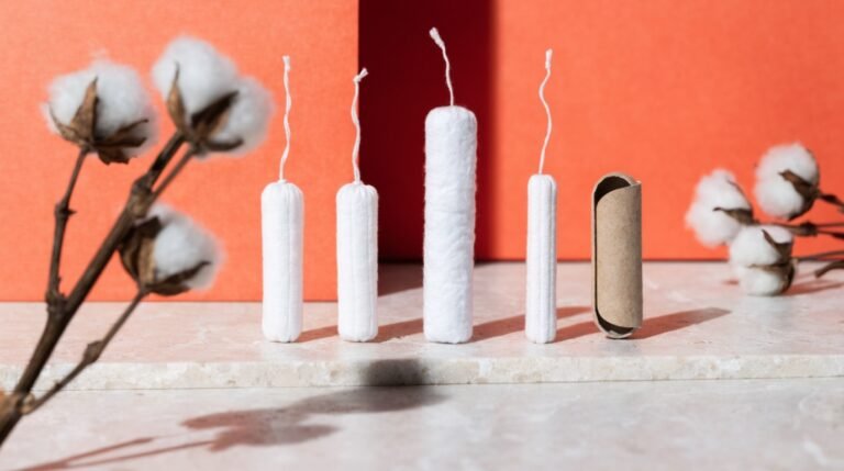 Unwrapped organic cotton tampons and a plain cardboard applicator arranged on a pale stone surface against a bold soft-coral background with dried cotton stems