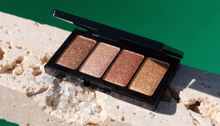 A shimmering pressed eyeshadow palette in warm bronze and champagne tones open on a pale stone ledge against a bold deep-green background with flakes of raw mica mineral catching the light