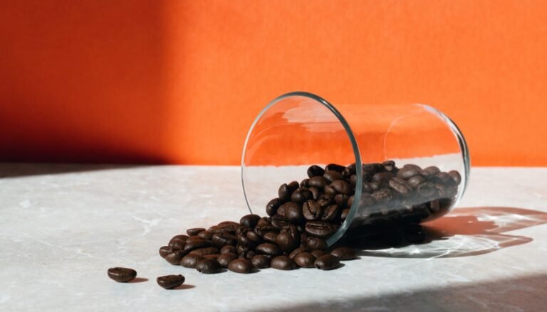 Roasted coffee beans spilling from a glass laboratory beaker across a pale stone surface against a bright citrus-orange background