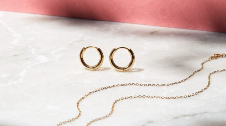 A pair of plain gold hoop earrings and a slim chain on a pale marble surface against a bold blush-pink background in soft directional daylight