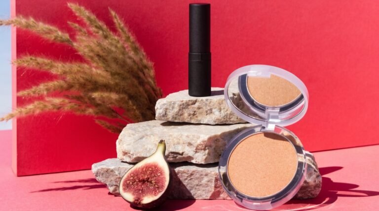 A black lipstick tube and an open compact of pressed powder raised on stacked stones against a bold coral-red background, with dried grasses and a halved fig in hard sunlight