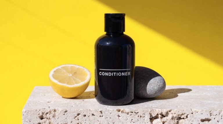 A glossy dark bottle labeled Conditioner on a travertine plinth against a bold sunny-yellow background with a halved lemon and a grey river stone in hard sunlight