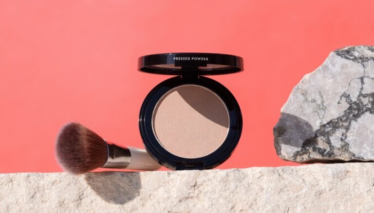 An open pressed powder compact and a soft makeup brush on a pale stone ledge against a bold coral background with a piece of raw grey mineral rock in bright sunlight