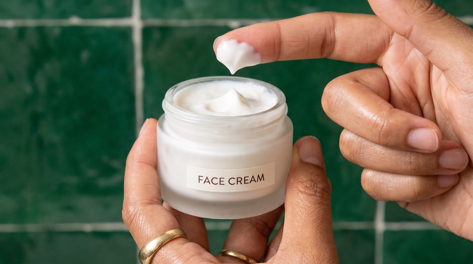 Two hands lifting a peak of white cream from an open frosted jar labeled face cream against a glossy green tiled wall